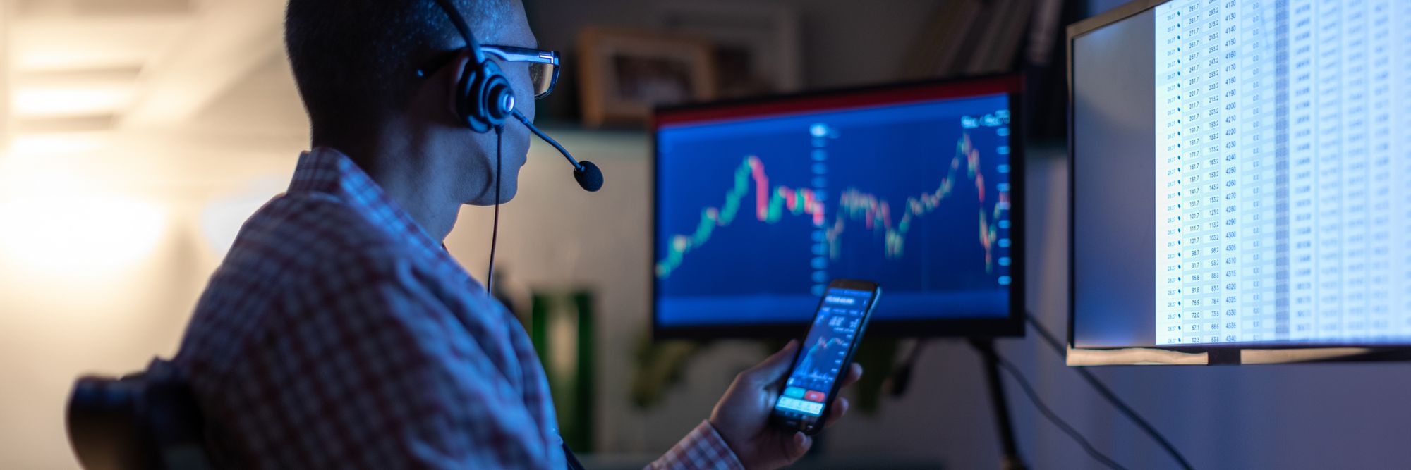 active investor at a computer screen, holding a phone, both showing stock charts. The trader is wearin a headset and sitting in the dark. 